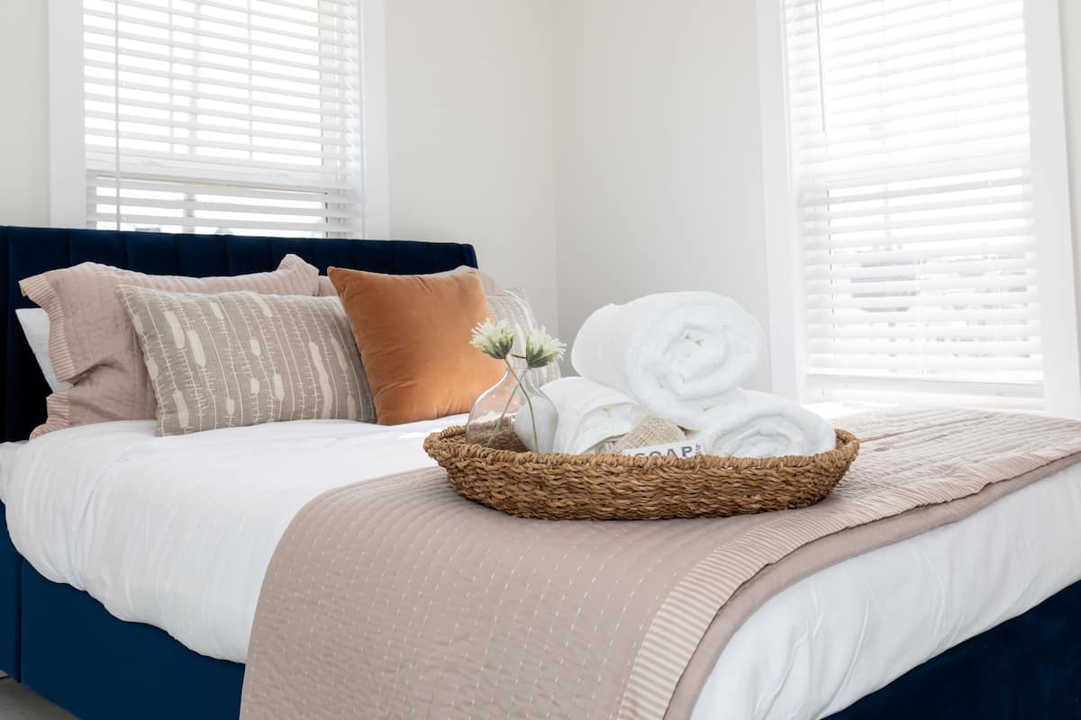 A neatly made bed with a blue headboard, a woven basket with folded towels, and a beige throw blanket.