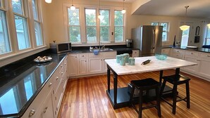 A modern kitchen with white cabinets, a black countertop, and a wooden floor. There are large windows providing natural light, a stainless steel refrigerator, and a dining area with two chairs.