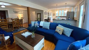 A spacious living room with a blue sectional sofa, wooden dining table, and chairs. A kitchen is visible in the background with modern appliances.