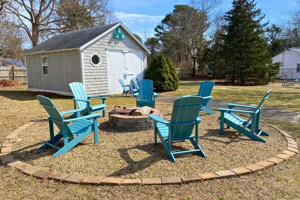 A small wooden shed with a circular seating area featuring four blue Adirondack chairs around a fire pit.