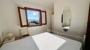 Un dormitorio con cama, una silla, una mesita de noche, una lámpara, una ventana con cortinas y un espejo.