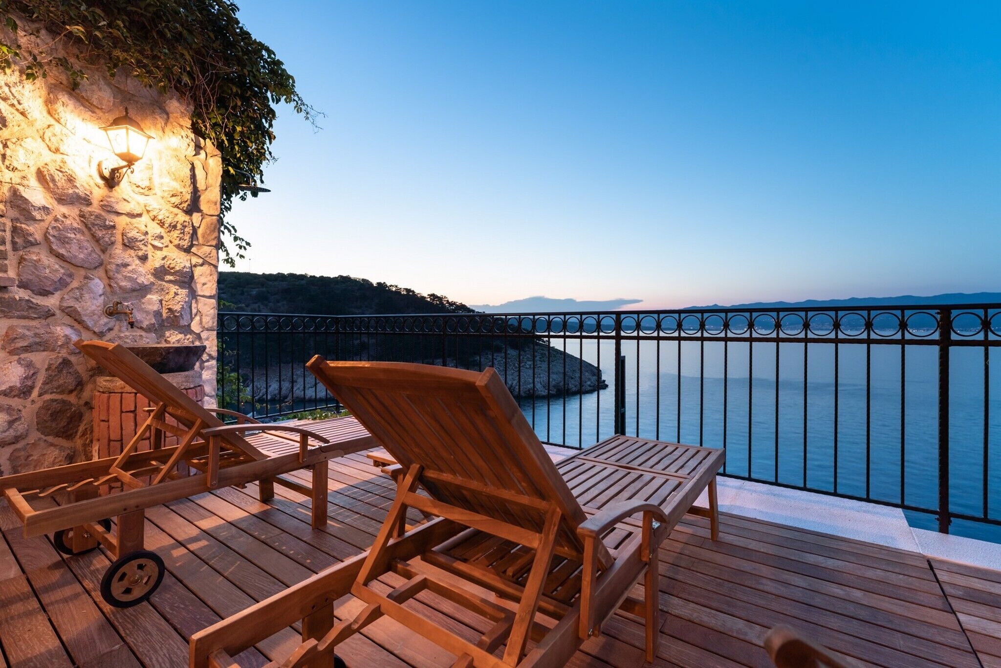 Una terraza de madera con dos sillas y una mesa, con vista a un cuerpo de agua y un muro de piedra con un farol.