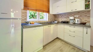 A modern kitchen with white cabinets, a Koncar refrigerator, a coffee maker, and a window with blinds. - Holiday apartment with terrace near the beach (Vrbnik)