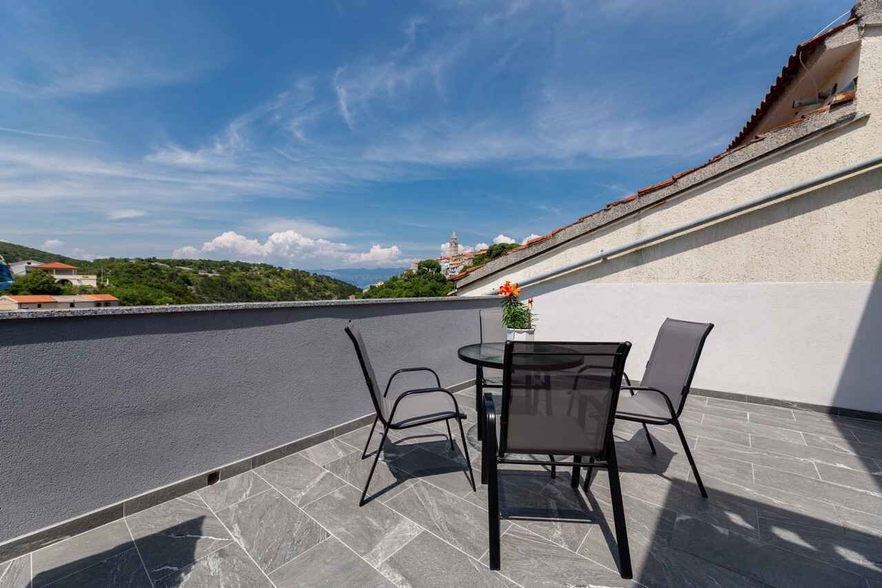 Eine Dachterrasse mit Tisch und Stühlen, einem Blumentopf und Blick auf einen Hügel mit Gebäuden.