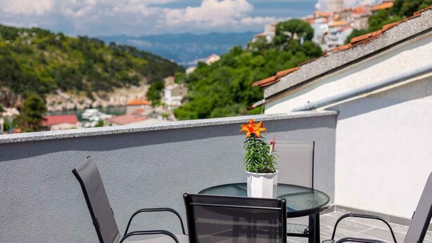 Une terrasse avec une table et des chaises, un pot de fleurs et une vue sur une ville avec ses bâtiments et une colline.