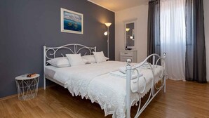 Une chambre avec un lit en métal blanc, une table d’appoint ronde, une table de chevet, une lampe fixée au mur, un tableau et une fenêtre avec des rideaux.