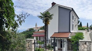 Une maison à deux étages, dotée d’un portail en pierre, d’un auvent rouge et blanc, et offrant une vue sur une ville avec un clocher en arrière-plan.