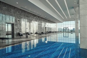 A modern indoor swimming pool with a long, narrow design, seating along the sides, and a large window providing a view of the cityscape. - ALSAWAH Aeon Towers - Premium 1BR with View Sheikh Zayed (6th of October City)