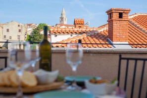 A terrace with a table set for dining, a bottle of wine, and glasses, overlooking a cityscape with traditional architecture and a tower. - Holiday home with balcony (Vrbnik)