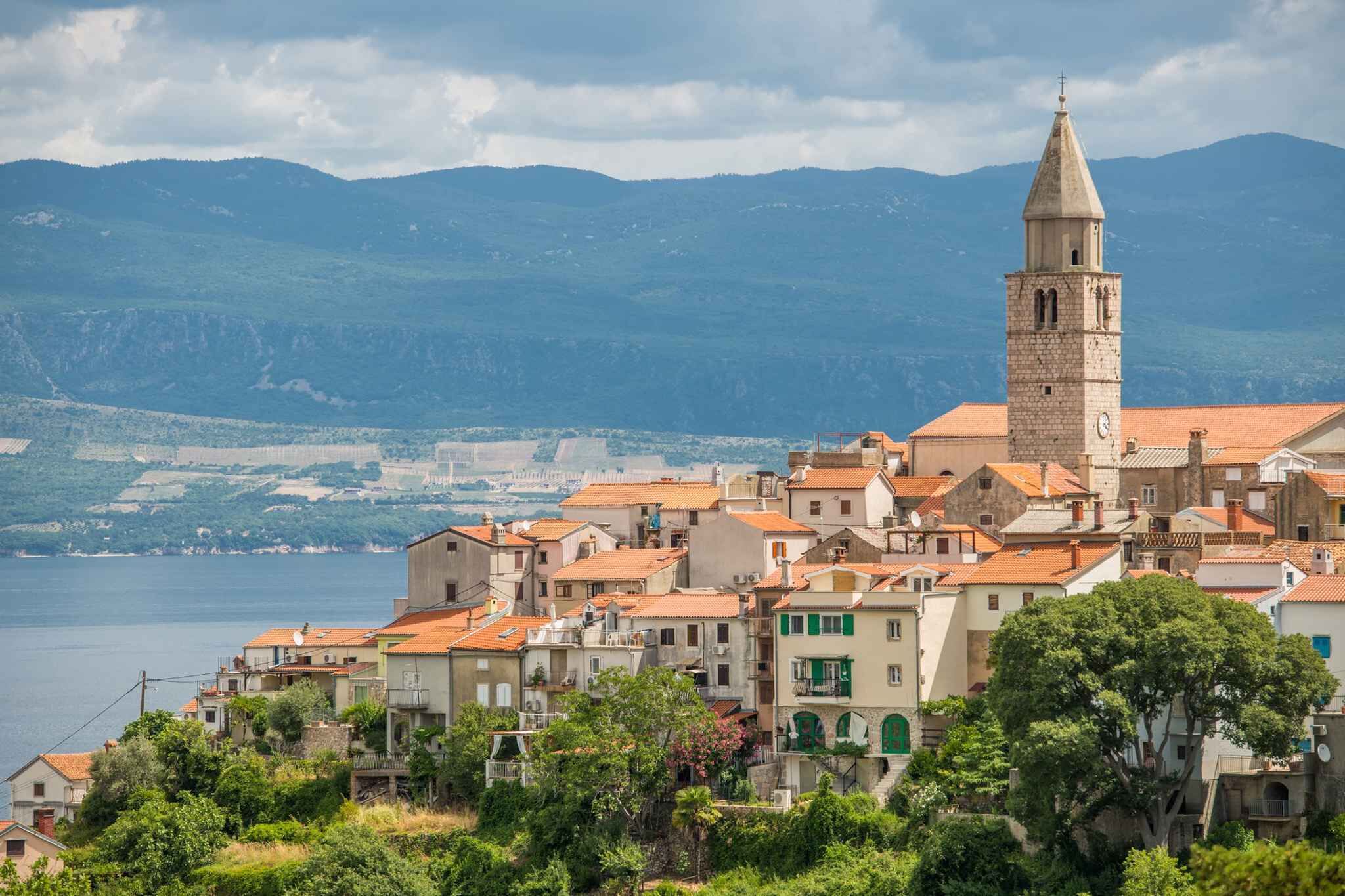 Eine Küstenstadt mit einem markanten Turm, terrassenförmig angeordneten Häusern und Blick auf das Meer und die Berge.
