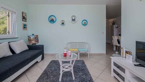 Ein Wohnzimmer mit einem dunklen Sofa, einem weißen Glastisch, einem weißen Korbstuhl und einer TV-Konsole.