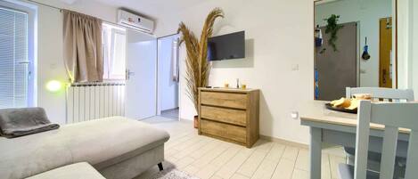 Une chambre moderne avec un canapé, une commode en bois, un téléviseur fixé au mur et une table à manger avec des chaises.
