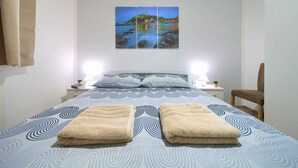 A neatly made bed with a patterned bedspread, two towels, a nightstand with a lamp, and a painting of a coastal scene above the bed.