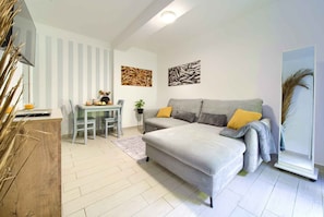 Uma sala de estar moderna com um sofá cinzento, uma pequena mesa de jantar e uma TV sobre um suporte de madeira.