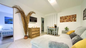 A modern living room with a sofa, a dining table, and a TV. There is a bed in the adjacent room.