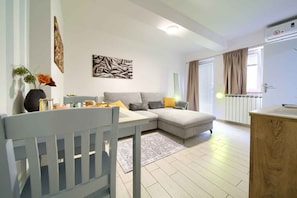 Uma sala de estar moderna com um sofá seccional cinza, uma mesa de jantar branca com cadeiras, uma mesinha lateral e um vaso decorativo com flores.