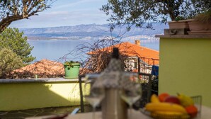 A terrace with a view of a lake and mountains, a table with a fruit basket, and a green plant.