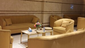 A spacious lounge area with a large beige sofa, a smaller armchair, and a round coffee table. The room features a high ceiling with recessed lighting and a marble floor. - Radwan Al Diyafah Hotel  (Makkah)