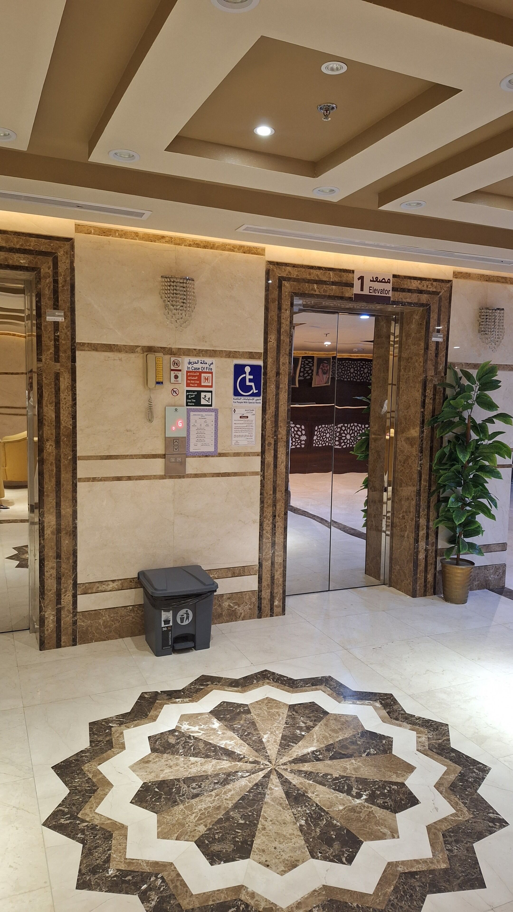 Entrada de ascensor con suelo de mármol, un cubo de basura y carteles informativos, incluyendo un letrero de emergencia contra incendios.