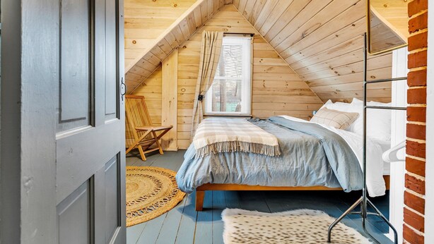 Ein gemütliches Schlafzimmer im Dachgeschoss mit schrägen Wänden, einem hölzernen Bett, einem Sessel und einem Fenster mit Vorhängen.