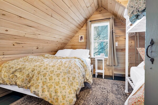 A cozy attic bedroom with a wooden sloped ceiling, a single bed with floral bedding, a small desk, and a window with blinds.