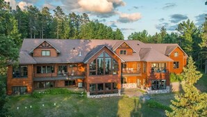 A large, multi-story log cabin with multiple balconies and large windows, surrounded by dense forest. - Bay Ridge North Lakeshore Lodge Retreat (Park Rapids)