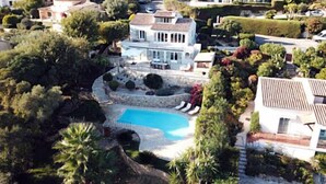 Una casa grande de varios pisos con piscina rodeada de exuberante vegetación y otras edificaciones residenciales.