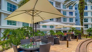 An outdoor patio area with wicker furniture and large umbrellas, surrounded by lush greenery and a multi-story building with balconies.
