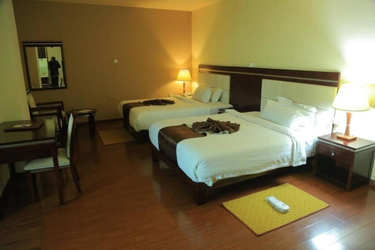 Une chambre d’hôtel avec deux lits, un bureau, une chaise, un miroir et une table de chevet avec une lampe.