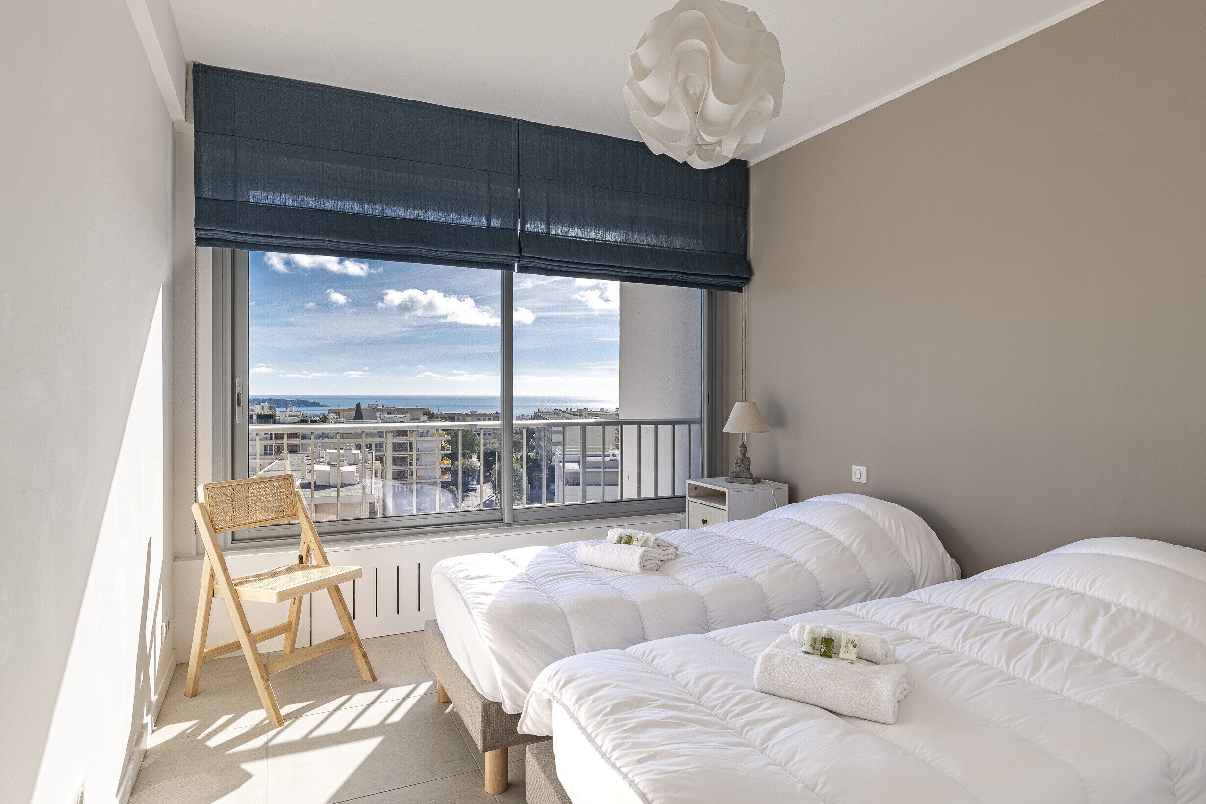 A bedroom with two beds, a chair, a nightstand, and a view of the city through large windows.