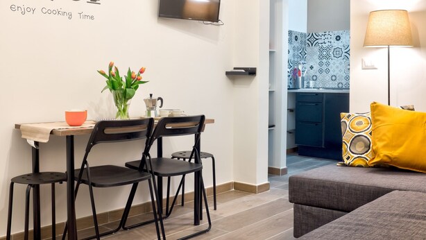Eine moderne Küche mit Essbereich, einem an der Wand montierten Fernseher und einem Sofa mit gelbem Kissen.