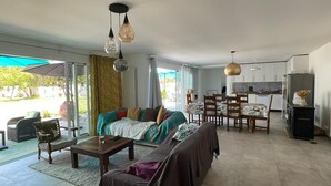 Un vaste espace de vie comprenant un canapé, une table basse et une table à manger. On y trouve également des luminaires suspendus et, au fond, un coin cuisine.