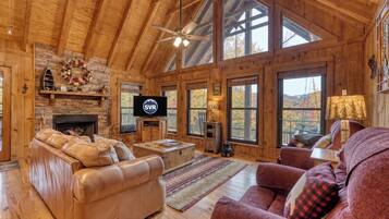 A spacious living room with a fireplace, leather sofas, a television, and large windows offering a view of the outdoors.