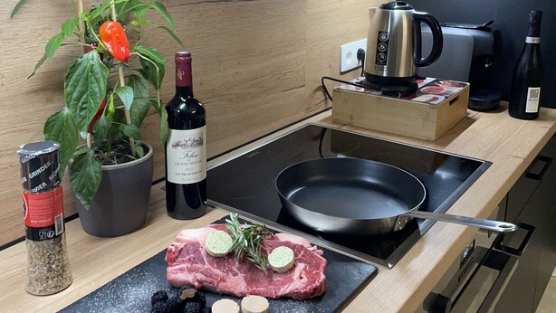 A modern kitchen with a wooden countertop, a kettle, a bottle of wine, a pan, and a cutting board with meat and herbs.