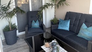 Een moderne buitenruimte met een glazen salontafel, twee banken met blauwe kussens en potplanten.