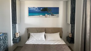 Een slaapkamer met een bed, twee kussens, een nachtkastje, een aan de muur hangend schilderij van een strand en een raam met gordijnen.