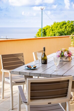 A wooden table with a bottle, glasses, and a plate of food on an outdoor patio with a view of the sea. - Arcobaleno Tropea (Tropea)