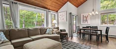 A modern living room with a sectional sofa, a dining table with chairs, and large windows offering a view of trees.