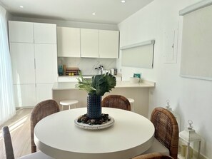 A modern kitchen with white cabinets, a round dining table with wicker chairs, and a large potted plant. - The Nest Garden Grove - by Zenbreak (Holetown)
