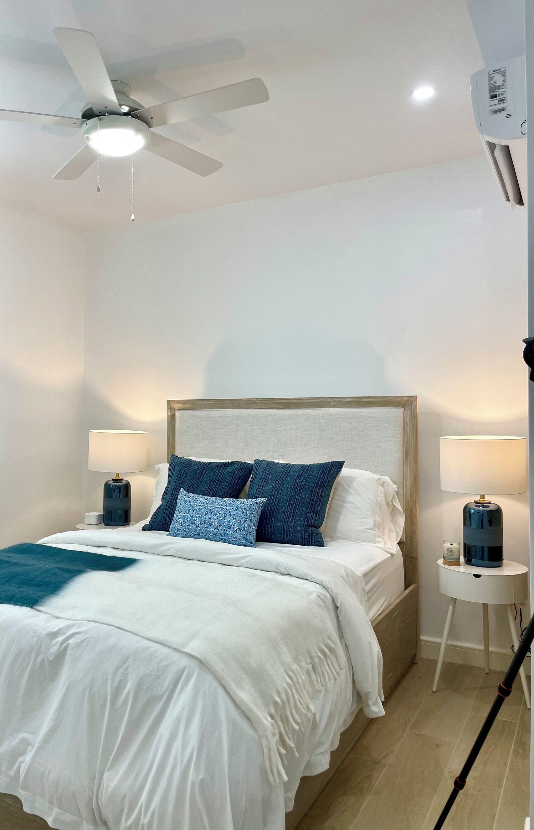 Un dormitorio ordenado con una cama, dos mesitas de noche con lámparas, un ventilador de techo y un equipo de aire acondicionado.