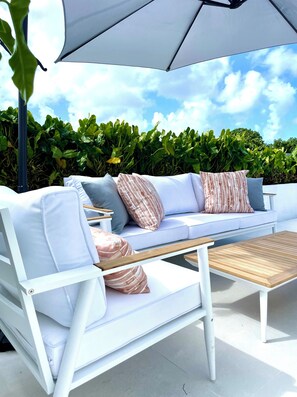 A white outdoor seating area with a sofa, cushions, and a wooden table. - The Nest Garden Grove - by Zenbreak (Holetown)