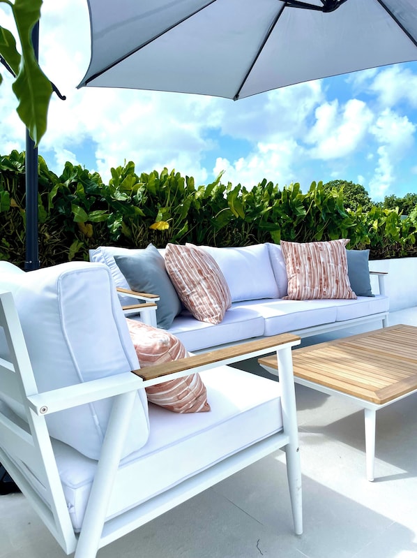 A white outdoor seating area with a sofa, cushions, and a wooden table.