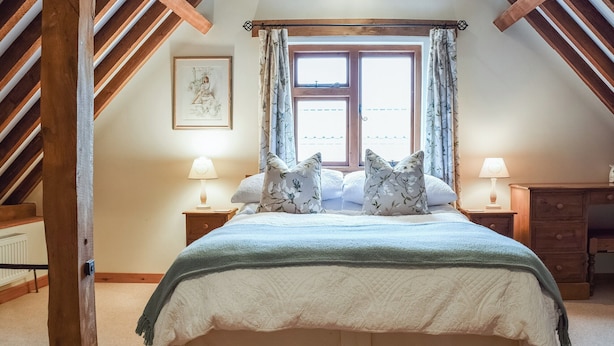 A bedroom with a large bed, two bedside tables with lamps, a window with curtains, and a wooden ceiling structure. - The Old Stable - Meadowbrook Farm (Moreton, near Thame)