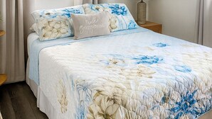 A bedroom with a floral bedspread, a bedside table with a lamp, a window with curtains, and a wall-mounted air conditioner. - Mauka Sunrise, Walk to Hau‘ula Beach & Trails, A/C (Hauula)