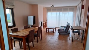 A living room with a wooden dining table, wicker chairs, a sofa, a TV, and a large window with sheer curtains.