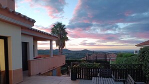 A patio with a wooden bench, a palm tree, and a view of distant buildings and hills under a pink and blue sky.