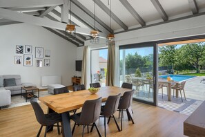 A modern living room with a wooden dining table, grey sofas, and a large window overlooking a swimming pool.
