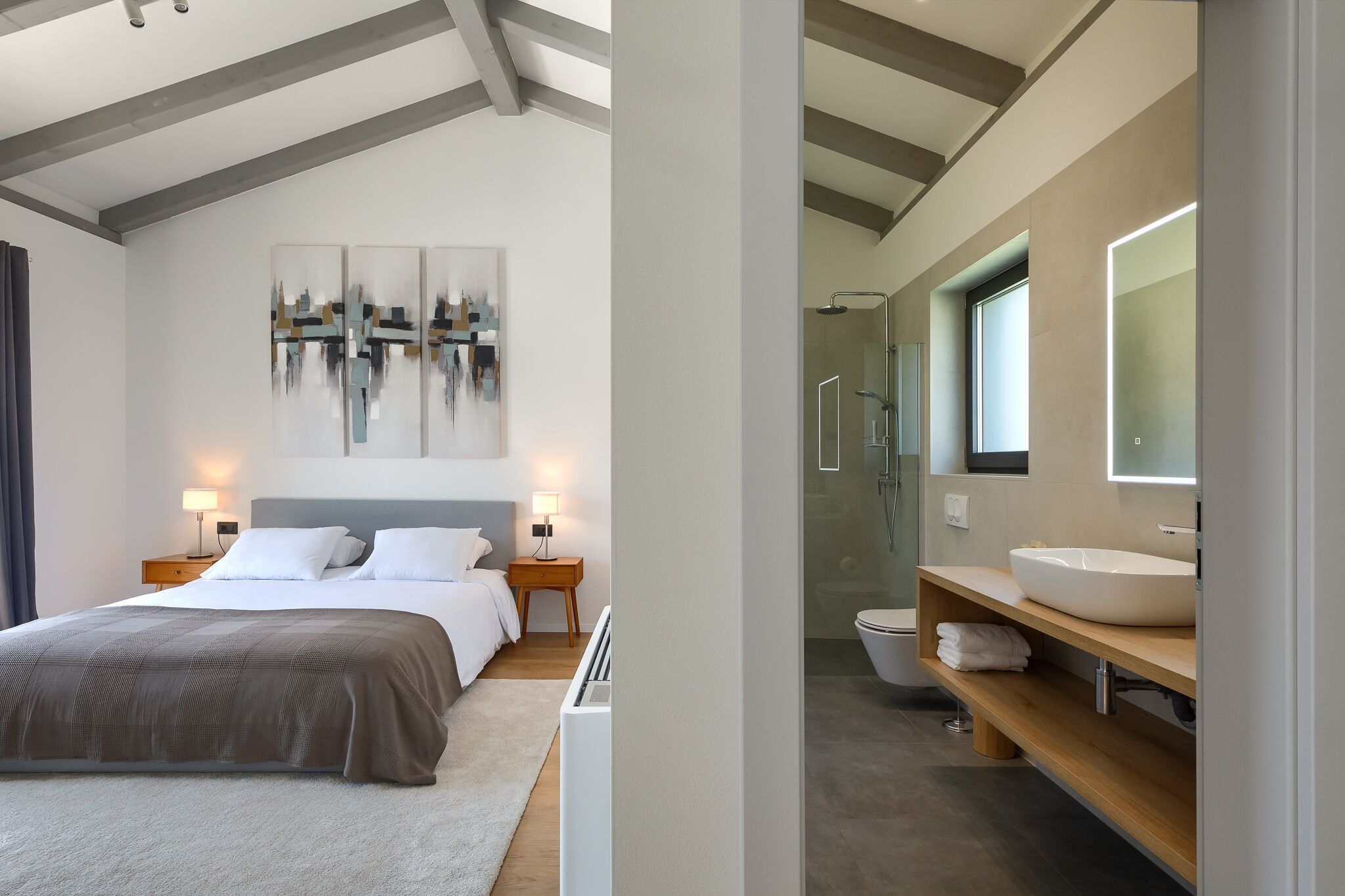 A modern hotel room with a large bed, bedside lamps, a wall-mounted abstract art piece, and a bathroom with a large mirror, sink, and shower.