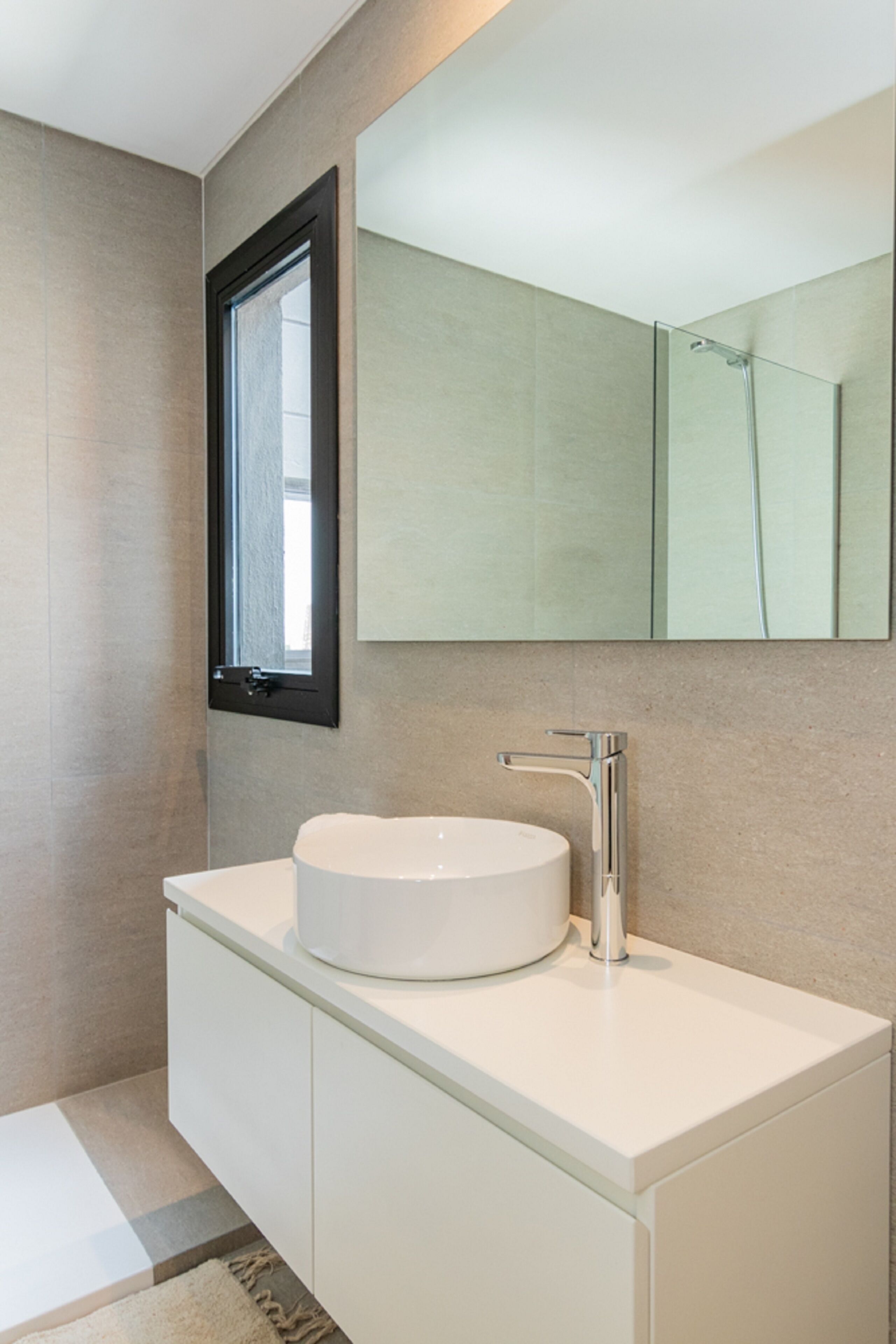 A modern bathroom with a round white sink, a large mirror, and a window.