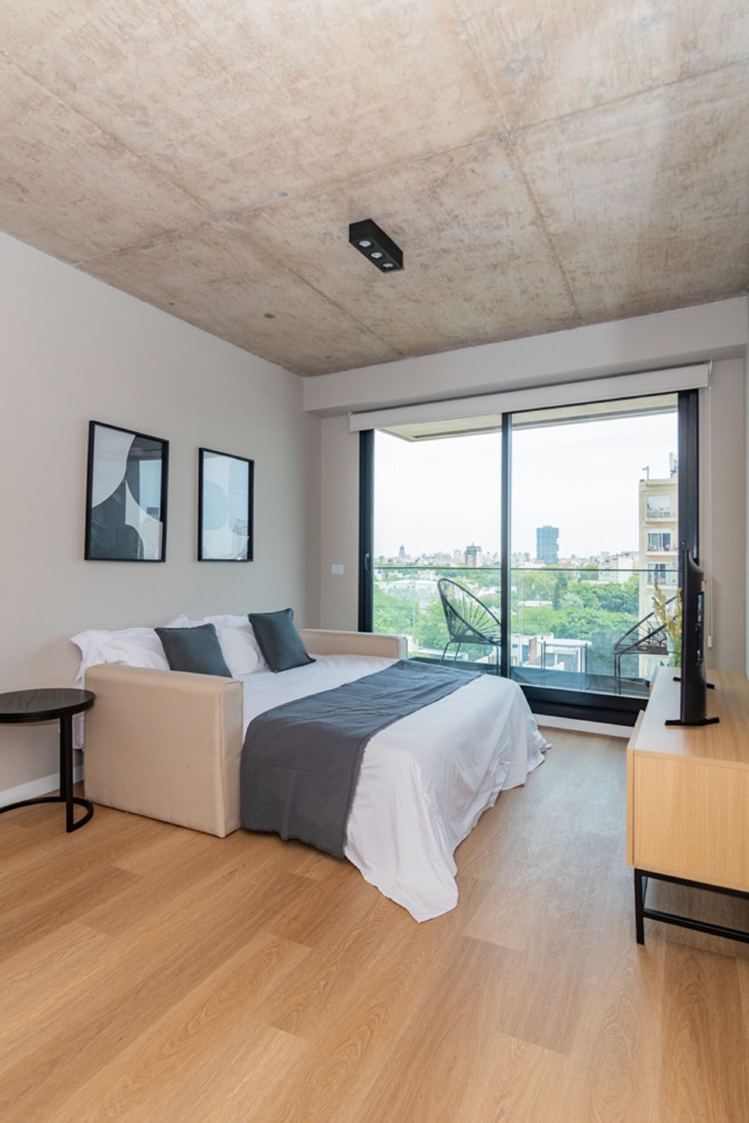 A modern bedroom with a large bed, a desk, and a view of the city through large windows.